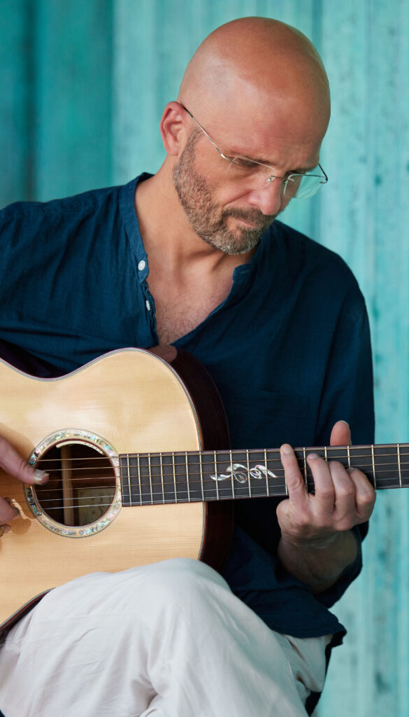 © Martin Moro / C&G Pictures Martin Moro sitzt mit Überschlagenen Beinen und spielt auf seiner Gitarre mit Blick auf den Gitarrenhals gerichtet.