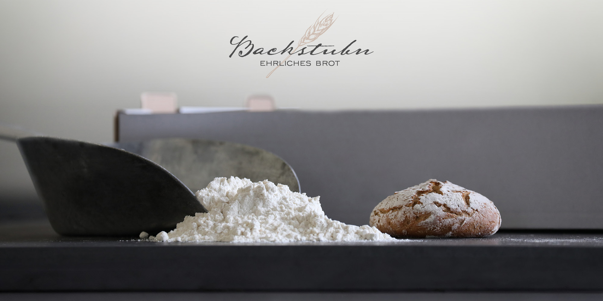 Foto © Simone Maurer-Jobst Das Logo „Backstubn – ehrliches Brot“ auf einem Foto von einer Schaufel mit Meh und einem kleinen Weckerl.