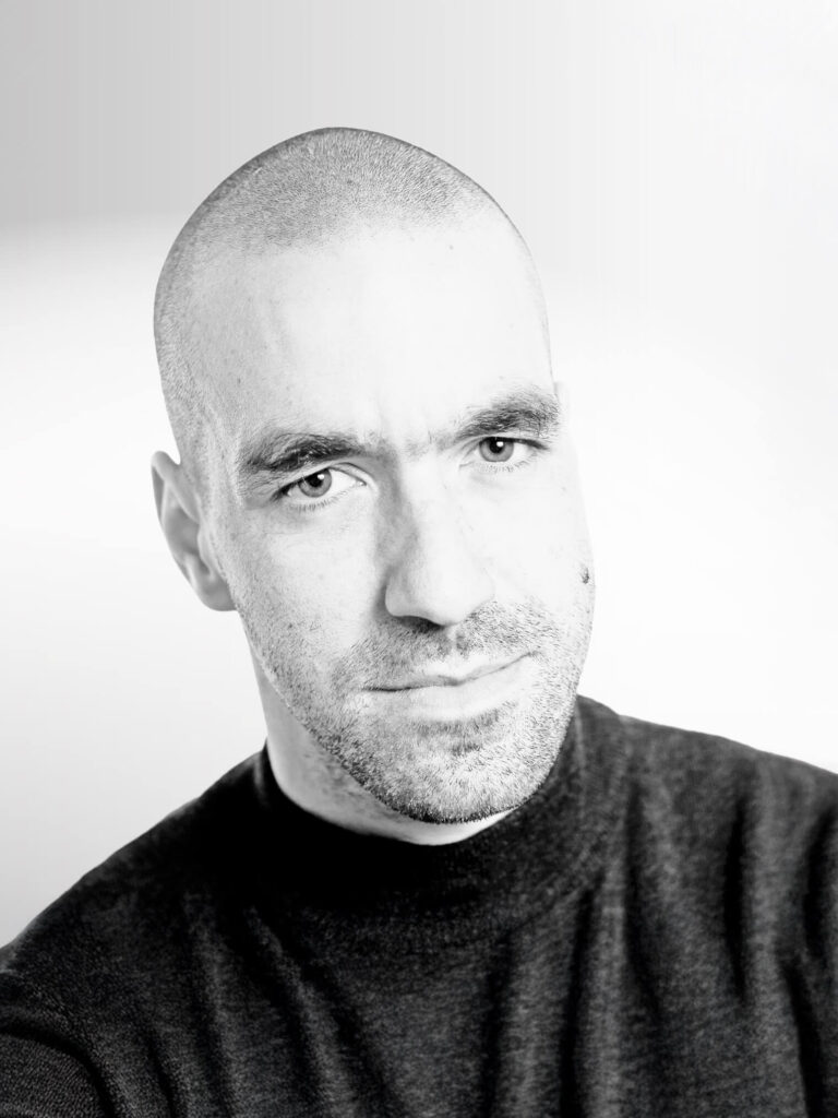 Martin Hinterdorfer: Schwarz-Weiß Porträtaufnahme eines Mannes mit rasiertem Kopf und Stoppelbart. Er trägt ein dunkles Oberteil. Der Blick ist direkt in die Kamera gerichtet.